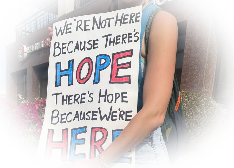 A woman holding a hand-painted sign reading "We're not here because there's hope, there's hope because we're here," with American flags nearby.